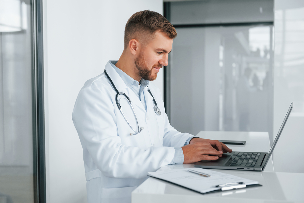 Usando laptop. Médico profissional de jaleco branco está na clínica.